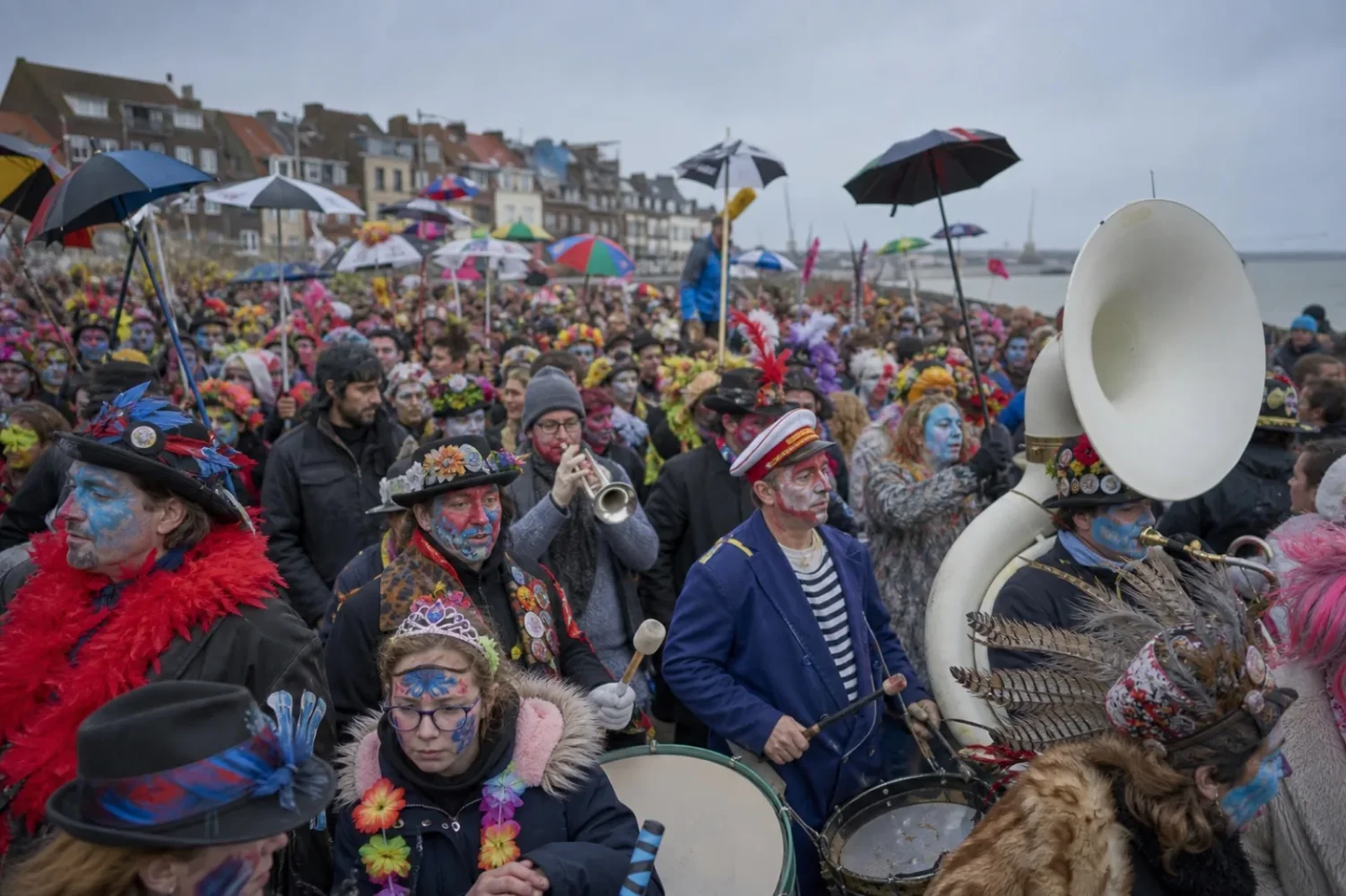 Foule costumée du carnaval de Dunkerque, avec parapluies, chapeaux à plumes et fanfare sous un ciel gris.