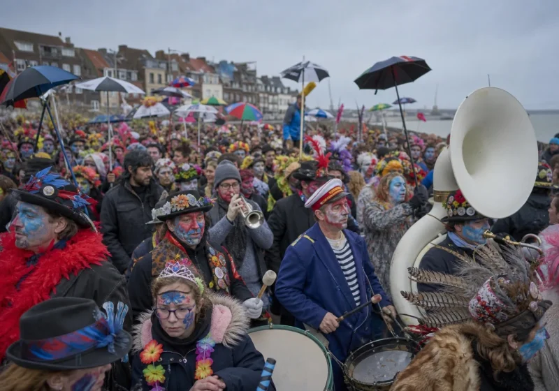 Foule costumée du carnaval de Dunkerque, avec parapluies, chapeaux à plumes et fanfare sous un ciel gris.