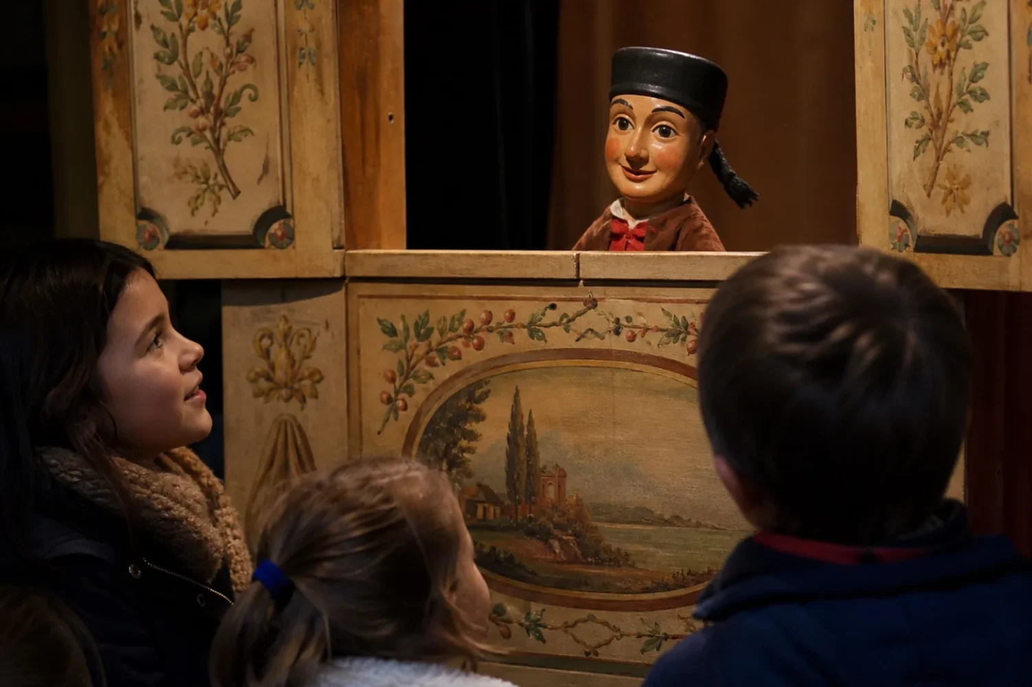 Marionnette de Guignol, avec son chapeau noir et sa tresse, dépassant du castelet devant des enfants.