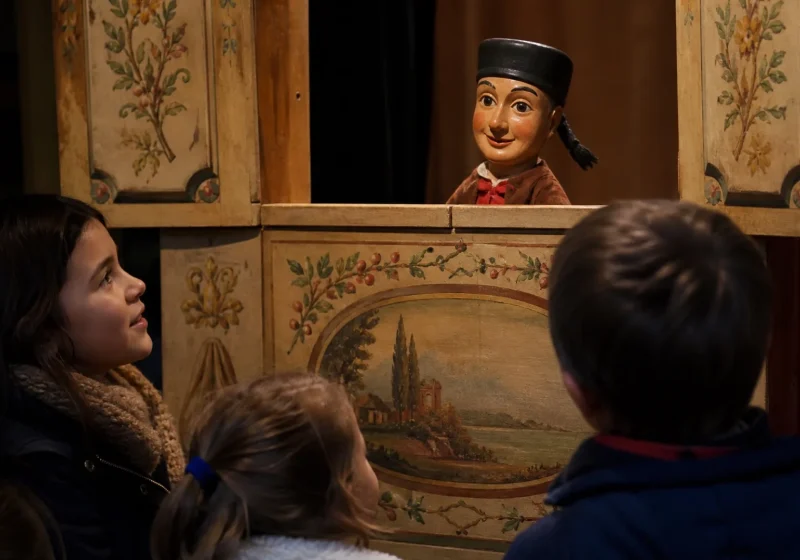 Marionnette de Guignol, avec son chapeau noir et sa tresse, dépassant du castelet devant des enfants.