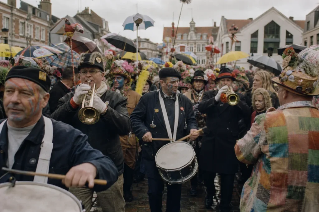 Fanfare de carnaval entraînant une foule dense sous un ciel gris, costumes bariolés et parapluies levés.