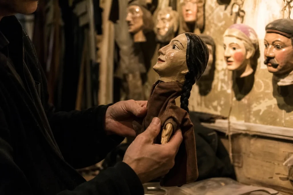 Coulisses d'un théâtre de marionnettes, mains tenant une marionnette à gaine de Guignol, têtes peintes au mur.